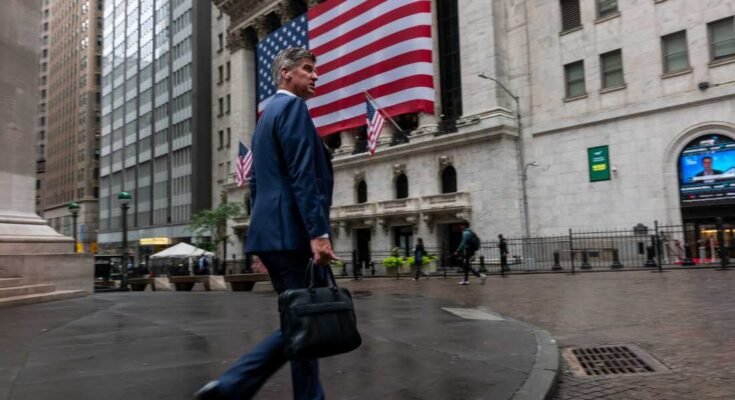 Egy sötétkék jelmezben lévő üzletember sétált a New York -i tőzsdén, amelynek homlokzatát hatalmas amerikai zászlóval borítják.
